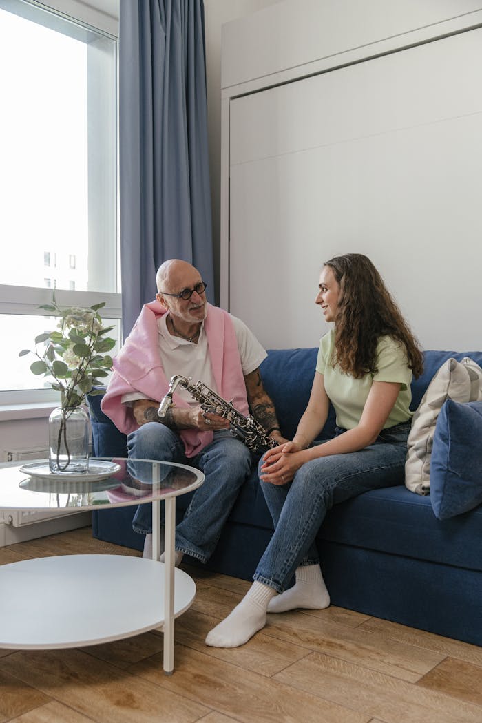 An elderly man instructs a young woman in saxophone playing on a sofa indoors.